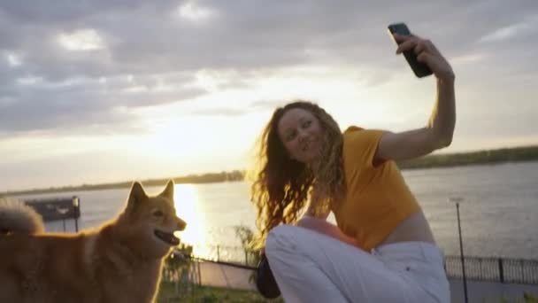 一个积极向上的女人在夕阳西下沿着堤岸散步的时候 带着她的红狗在智能手机上自拍 一个女孩和她的四条腿的朋友呆在一起 — 图库视频影像