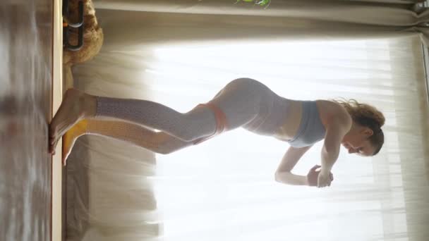 一个女人在家里用橡胶在一个运动垫上训练她的胶状肌肉蹲 一个垂直的小腿 一位年轻的运动女子正在做家庭运动 一只红头发的猫在后台玩耍 — 图库视频影像