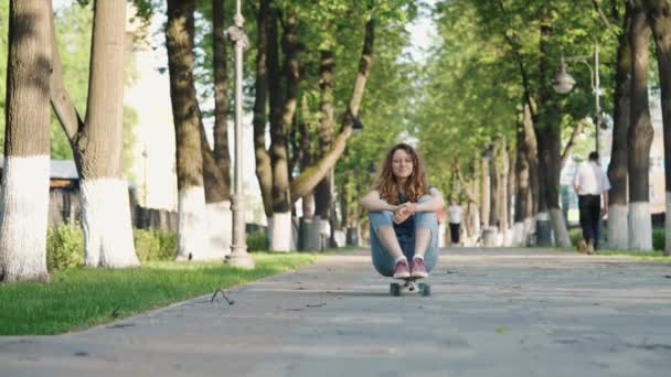 可爱的无忧无虑的女孩抱着胳膊坐在滑板上沿着小巷滚下山 在夏天的公园里散步休息 一个年轻的女人做积极的运动很开心 — 图库视频影像