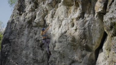 Dişi bir kaya tırmanışçısı, dik bir kaya boyunca koruyucu bir miğferle iple bir rotaya tırmanır. Tırmanma ayakkabısı giyen bir kız ayak basacak yer arıyor ve yorgun ellerini sallıyor..