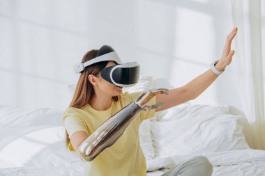 Bir kadın sanal gerçekliği keşfediyor, bir VR kulaklık takıyor ve parlak bir yatak odasında fütürist bir robotik kolla etkileşime giriyor, gelişmiş teknolojiyle sanal deneyimlerin karışımını simgeliyor. Kadın