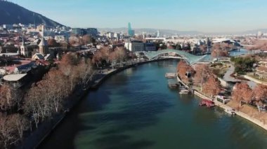 Açık mavi bir gökyüzü altında çıplak ağaçlar ve mimari simgeler sergilenen kışın nehir kenarındaki bir şehir manzarasının panoramik bir görüntüsü. Gürcistan yakınlarındaki Tiflis 'teki Kura Nehri' nin havadan görüntüsü.