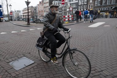 Sakallı, şapka ve ceket giyen yaşlı bir adam, Avrupa 'da bir şehir merkezinde kaldırım taşı caddesinde bisiklet sürüyor. Hollanda, Amsterdam, Ocak 2020.