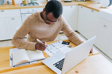 Ev Planlarını Laptop ve Hesap Makinesinin yanına Odaklanmış Afro-Amerikan Adam Analiz Ediyor. Gözlüklü nişanlı bir adam mutfak masasının mimari planlarını gözden geçirmeye odaklanmış..