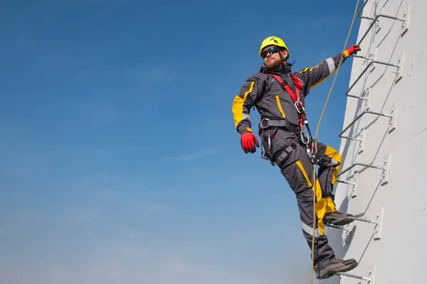 Industrial climber, a worker in a helmet and a safety belt, holds on to the ladder with one hand, fastened to a climbing rope, looks into the distance against the blue sky, copyspace. Carrying out