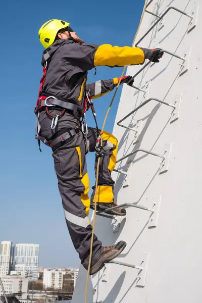 A male industrial climber climbs up a metal ladder in safety gear and a helmet to perform installation work. Carrying out high-rise works on metal structures.