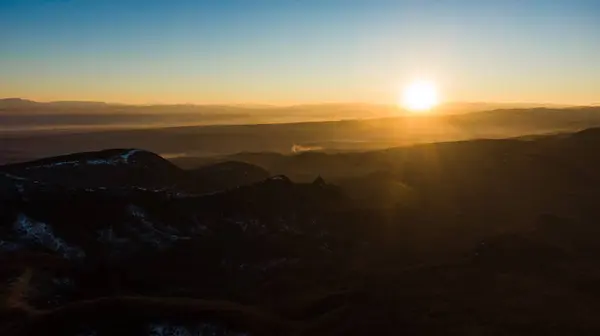 The sun rises over a serene mountain landscape, casting a warm glow over the misty valleys and undulating hills, captured in an expansive aerial view.
