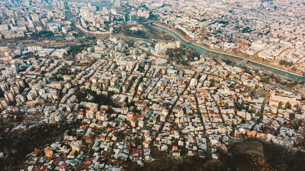 A sprawling urban tapestry, this aerial shot captures the dense clustering of buildings and the serpentine flow of a river through the heart of a bustling city. Aerial view of Tbilisi at sunset, dense