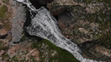 Güçlü bir şelalenin tepeden aşağı görüntüsü engebeli araziyle çevrili, altında ölçekli insanlar var. Doğanın gücü, bu çarpıcı sahnede insan varlığıyla çelişiyor..