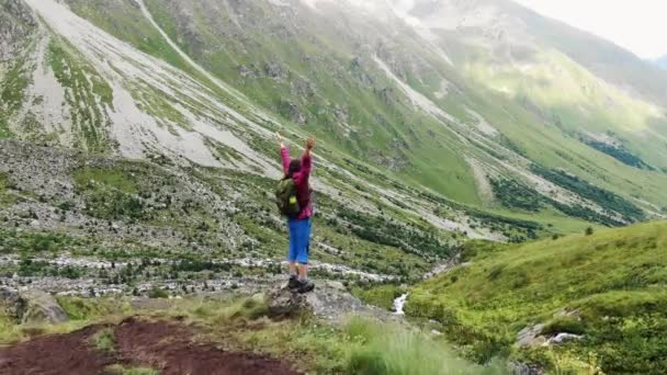 勝利で育った腕を持つハイカーの女性は 冒険と達成の精神を体現し 山の谷の広大な広がりを眺めながら岩の上に立っています — ストック動画