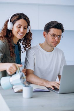 Gülümseyen bir kadın, erkek arkadaşı dost canlısı ve üretken bir ofis ortamı olan dizüstü bilgisayara odaklanırken kahve koyuyor..