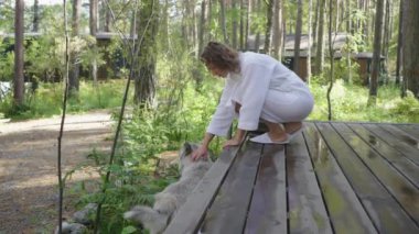 Beyaz önlüklü bir kadın, bir orman kulübesinin verandasında bir köpeği okşadı. Sabah, glamping evinde. Çam ormanlarında kır tatili.