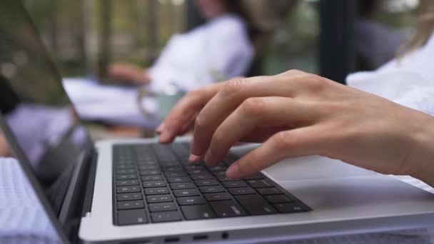 在笔记本电脑键盘上工作的女性手指的特写 两名女同事在网上的一台电脑上工作 她们坐在树林里的一个木制平台上喝咖啡 在一个国家的远程工作 — 图库视频影像
