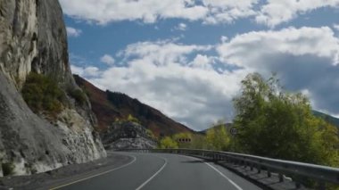 Dağ zirveleri ve dinamik bulutlarla dolu dramatik bir kaya yüzeyinin yanındaki dolambaçlı bir yolun büyüleyici görüntüsü bir yol gezisinde doğanın ihtişamını anımsatıyor. Birinci şahıs görünümü.