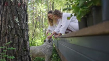 Sıcak beyaz bornozlu iki arkadaş, ahşap bir güvertede yumuşacık bir köpekle ormanın yemyeşil bir bölgesinde nazik bir anın tadını çıkarıyorlar. Kızlar kendine gelen yerel bir köpeği okşuyor.