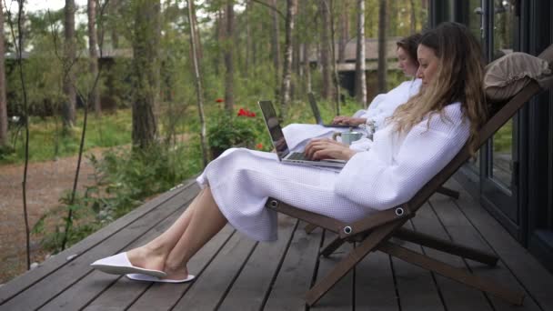 两名女自由职业者穿着白色外套 坐在森林中央一座乡村房屋的木制阳台上的椅子上 在笔记本电脑上工作 在松木中舒适的房子里上网工作 — 图库视频影像
