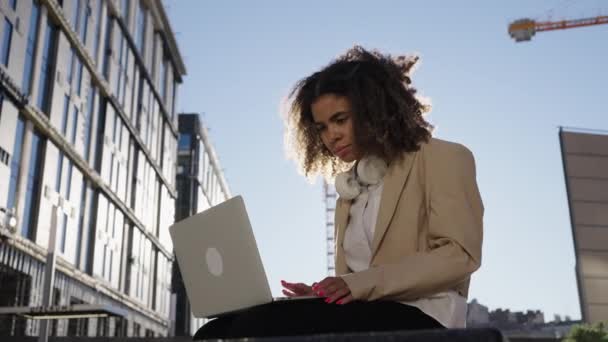 非洲裔美国女商人坐在商业中心附近的长椅上 在阳光的映衬下 在笔记本电脑前在线工作 身穿夹克衫的女自由职业者做生意 — 图库视频影像
