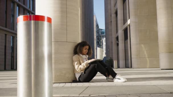 今年夏天 非裔美国女自由职业者学生靠着一根柱子 坐在一栋现代写字楼附近的水泥地上工作 这就是远程办公和学习的概念 — 图库视频影像