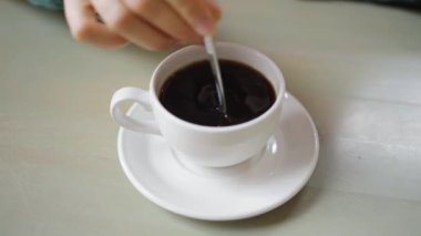Bir insan çay tabağında beyaz seramik bardakta dumanı tüten bir fincan kahveyi karıştırır. Beyaz Kupa 'da karıştırılmış taze siyah kahve 