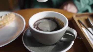 Bir el, bir fincan sade kahvenin içine süt dökerek, yüzeyde dönen bir desen oluşturur, rahat bir kafe ortamında kahvaltı düzeniyle birlikte. Seramik 'te sütlü kahvenin içine krema dök