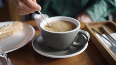 Bir insanın taze demlenmiş kahveye krema karıştırıp seramik bardaktaki dokuyu ve hareketi vurguladığı yakın plan bir fotoğraf. Bir kadının eli bir fincan kahve alır ve aromasının tadını çıkarır.