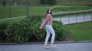 Kulaklıklı ve sırt çantalı genç bir kadın şehir parkında neşeli bir şekilde yürüyor, müziğinin keyfini çıkarıyor ve güneşli bir yaz gününün coşkulu atmosferini kucaklıyor.