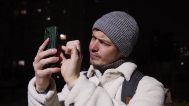 Beyaz ceketli ve bereli genç gezgin, gece manzarasını yakalıyor, dijital fotoğraf için akıllı telefonu kullanıyor ve kış akşamları şehir ortamını keşfediyor.