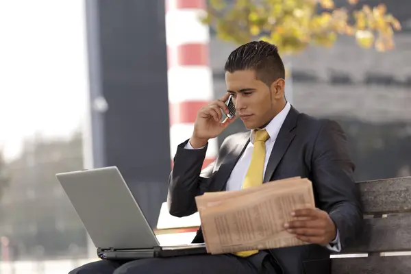 ビジネスマンはラップトップと仕事をするベンチに座り 携帯電話で電話して新聞を読んだ ストックフォト