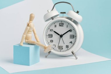 Tahta adam beyaz bir çalar saatin yanında oturuyor. Zamanı izleme kavramı, uzun süre bekle..