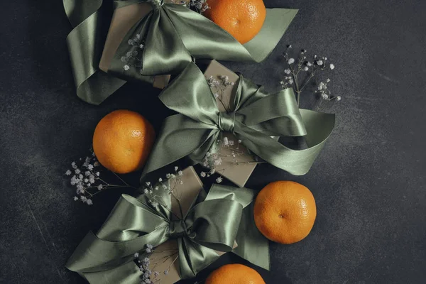 Gift boxes with olive green ribbons tied in a bow, tangerines, black background, top view. Decoration for holidays, birthdays, weddings.