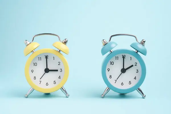 Two alarm clocks of blue and yellow colors, the hands show two o\'clock and three o\'clock. Concept of winter and summer time.