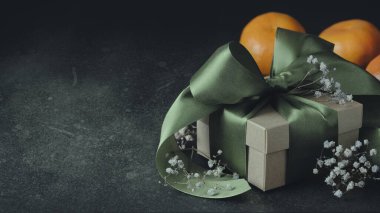 Yeşil kurdeleli hediye kutusu, fiyonk, mandalina ve küçük çiçekler. Siyah arka plan, üst manzara. Solda mesaj için yer var. Tatiller, doğum günleri, düğünler için dekorasyon.