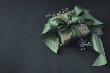 Yaya bağlanmış zeytin yeşili kurdeleli kağıt hediye kutusu, küçük çiçekler, siyah arka plan. Solda mesaj için yer var. Tatiller, doğum günleri, düğünler için dekorasyon.