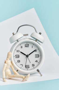 Tahta adam beyaz bir çalar saatin yanında oturuyor. Geç kalma kavramı, uzun bekleyiş, zamanın geçişi..