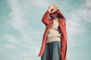 Kırmızı paltolu ve şapkalı genç kız gökyüzüne karşı duruyor. Düşük açı görünümü.