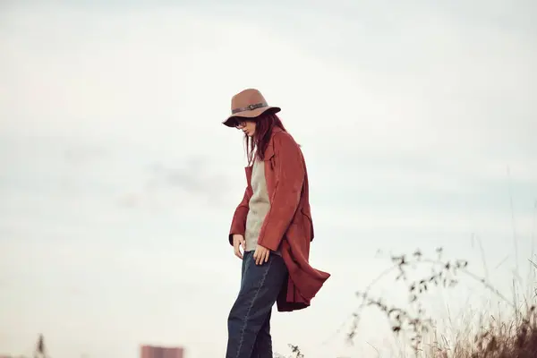 stock image Girl in hat and red coat stands against the sky, grass in the foreground.