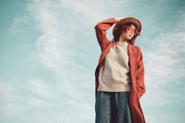 Kırmızı paltolu ve şapkalı genç kız gökyüzüne karşı duruyor. Düşük açı görünümü.