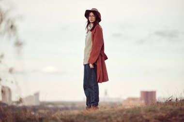 Sakin, kendine güvenen, şapkalı ve kırmızı paltolu bir kız kuru çimlerin üzerinde duruyor gökyüzünün ve şehrin arka planında ve kameraya bakıyor..