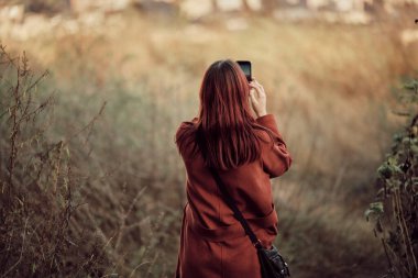 Kırmızı ceketli, uzun kızıl saçlı bir kız sonbahar otlarının arasında dikiliyor ve akıllı telefonuyla manzara fotoğrafı çekiyor..