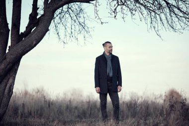 Sonbahar ve kış mevsimlerinde bir ağacın yanında duran mavi ceketli ve yün atkılı sakallı orta yaşlı bir adamın portresi..