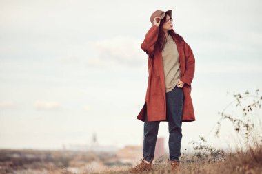 Kızıl saçlı, uzun saçlı genç kız elinde şapkasıyla gökyüzü ve şehir manzarasının arka planında çimlerin üzerinde duruyor..