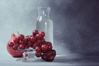 Su sürahisi, nar ve kırmızı üzümler cam tezgahta, desenli gri arkaplan