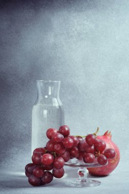 Su sürahisi, nar ve kırmızı üzümler cam tezgahta, desenli gri arkaplan