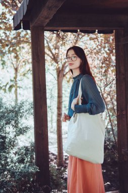 Tahta bahçe kemerinde çantalı genç bir kız, yeşilliklerle çevrili duruyor. Tarz sahibi ve çevre dostu bir portre. Sürdürülebilir yaşam ve doğa bağlantısı kavramı.