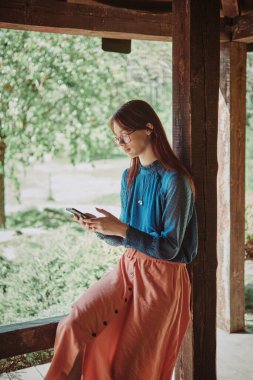 Kırsal ahşap bir çardakta akıllı telefon kullanan genç bir kız doğanın ortasında teknolojiye gömülmüş. Dijital bağlantıya odaklı sıradan bir yaşam tarzı portresi. Dinginlik ve dijital çağ kavramı.