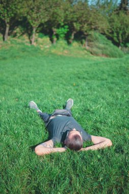 Kolları başının arkasında çimenlerde yatan bir adam. Park ortamında açık hava fotoğrafçılığı. Rahatlama ve doğa kavramı.