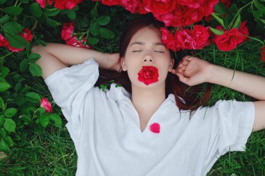 Beyaz gömlekli genç bir kadın çimenlerin üzerinde yatıyordu. Etrafı kıpkırmızı güllerle doluydu. Gözleri kapalıydı ve ağzında bir gül vardı..