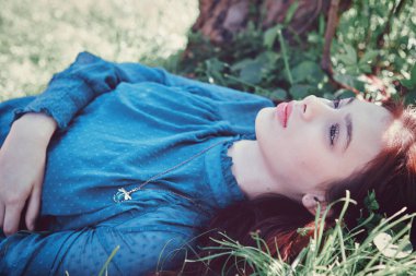 Çimlerin üzerinde rahatlayan genç kız, yakın plan. Açık havada sıradan bir bahar günü, yeşillik ve çiçeklerle doğal ortam. Huzurlu gençlik portresi.