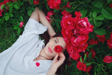 Beyaz gömlekli genç bir kadın çimenlerin üzerinde kırmızı güllerle uzanmış, bir gözünü kaplayan bir gülle gülümsüyor, neşe taşıyor ve doğayla derin bir bağ kuruyor..
