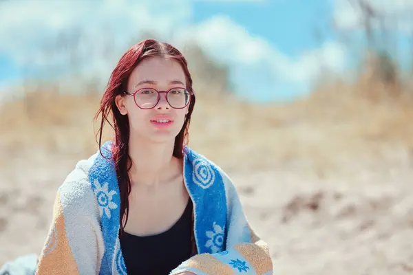 stock image Teenage girl with glasses wrapped in a towel, sitting on a beach with a distant look, surrounded by a sunlit sandy background, conveying a relaxed and contemplative summer mood.